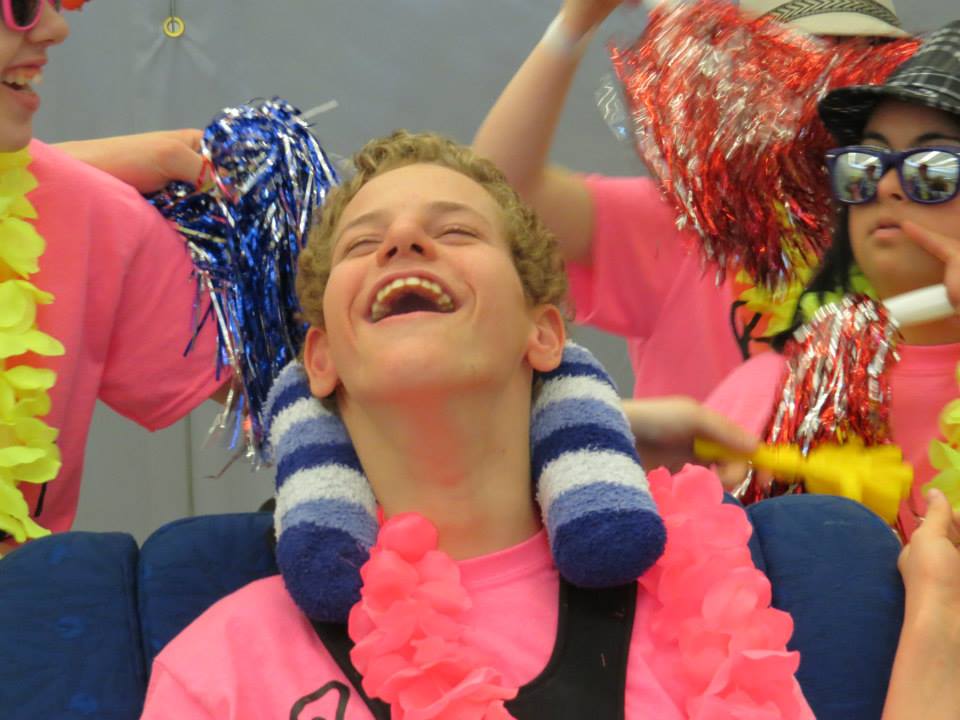 Laura MacGregor's son Matthew, looking up and smiling joyfully with eyes closed, 3 other youths standing behind him. All are sporting pink shirts, pompoms and flowered necklaces.