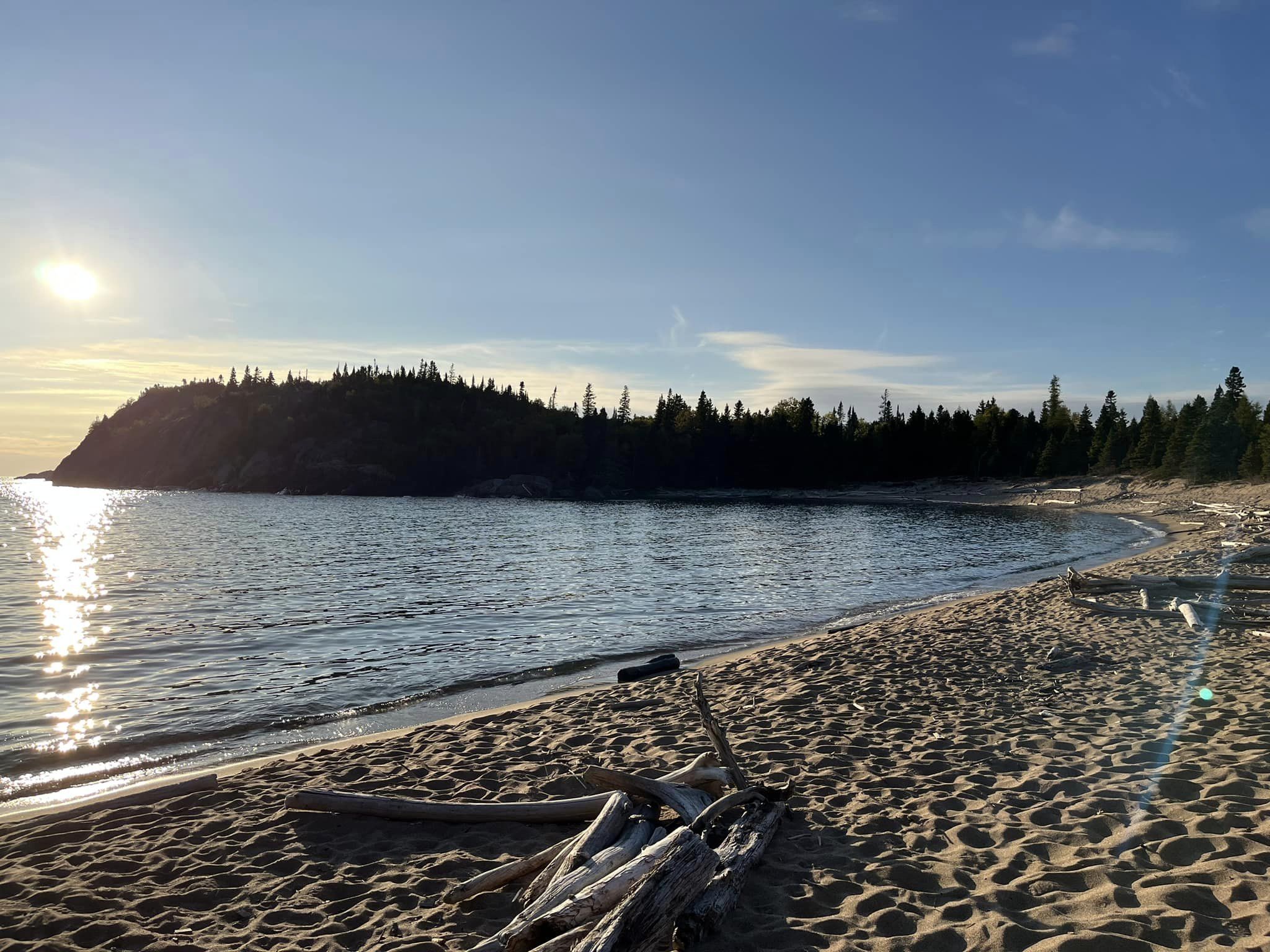 A sandy beach on Lake Superior; the sun is low and the lake is reflecting the very blue sky and bright gold sunlight. There are silhouettes of trees on the bank in the distance.