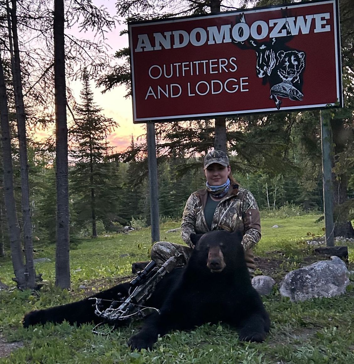 a woman in camouflage hunting gear poses with her bow and a bear she shot, in a green forest in front of a pink sunset. A red sign above reads 'Andomoozwe Outfitters and Lodge'.