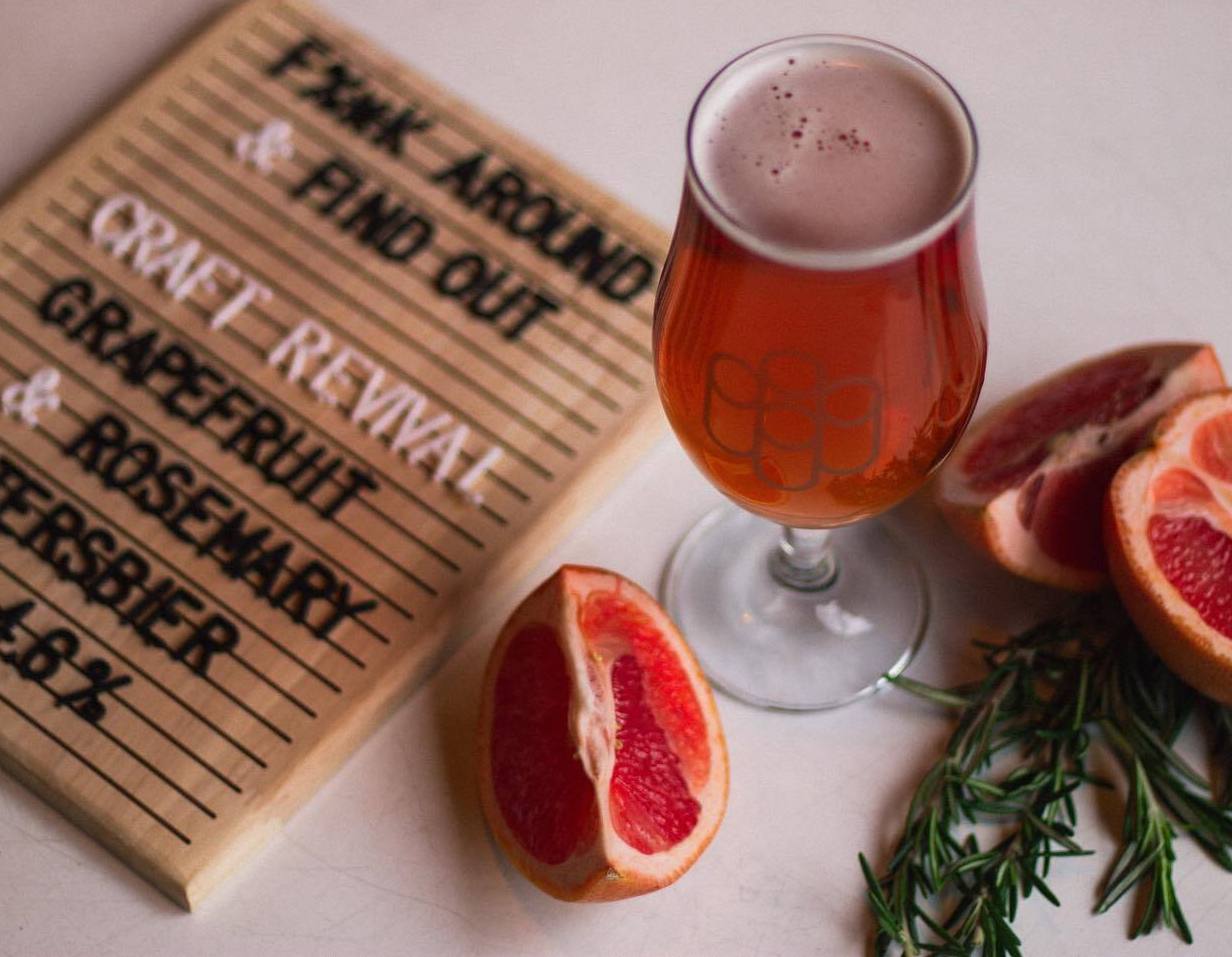 a glass of beer, some grapefruit wedges and fresh rosemary, next to a signboard that says "Craft Revival Grapefruit & Rosemary Patersbier 4.8%"