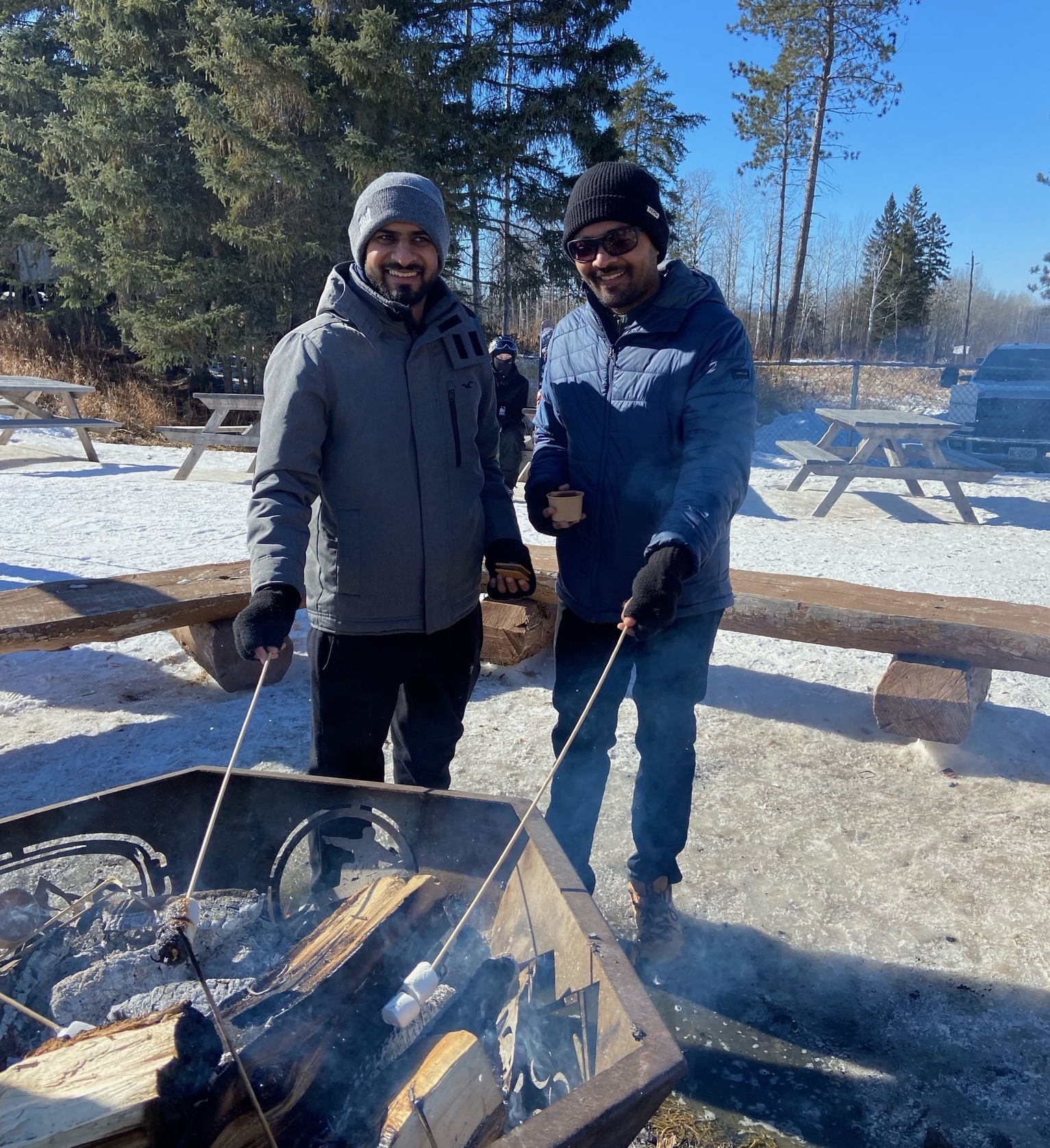 two men wearing winter outdoor clothing, smiling as they roast marshmallows over an outdoor fire on a sunny winter's day.
