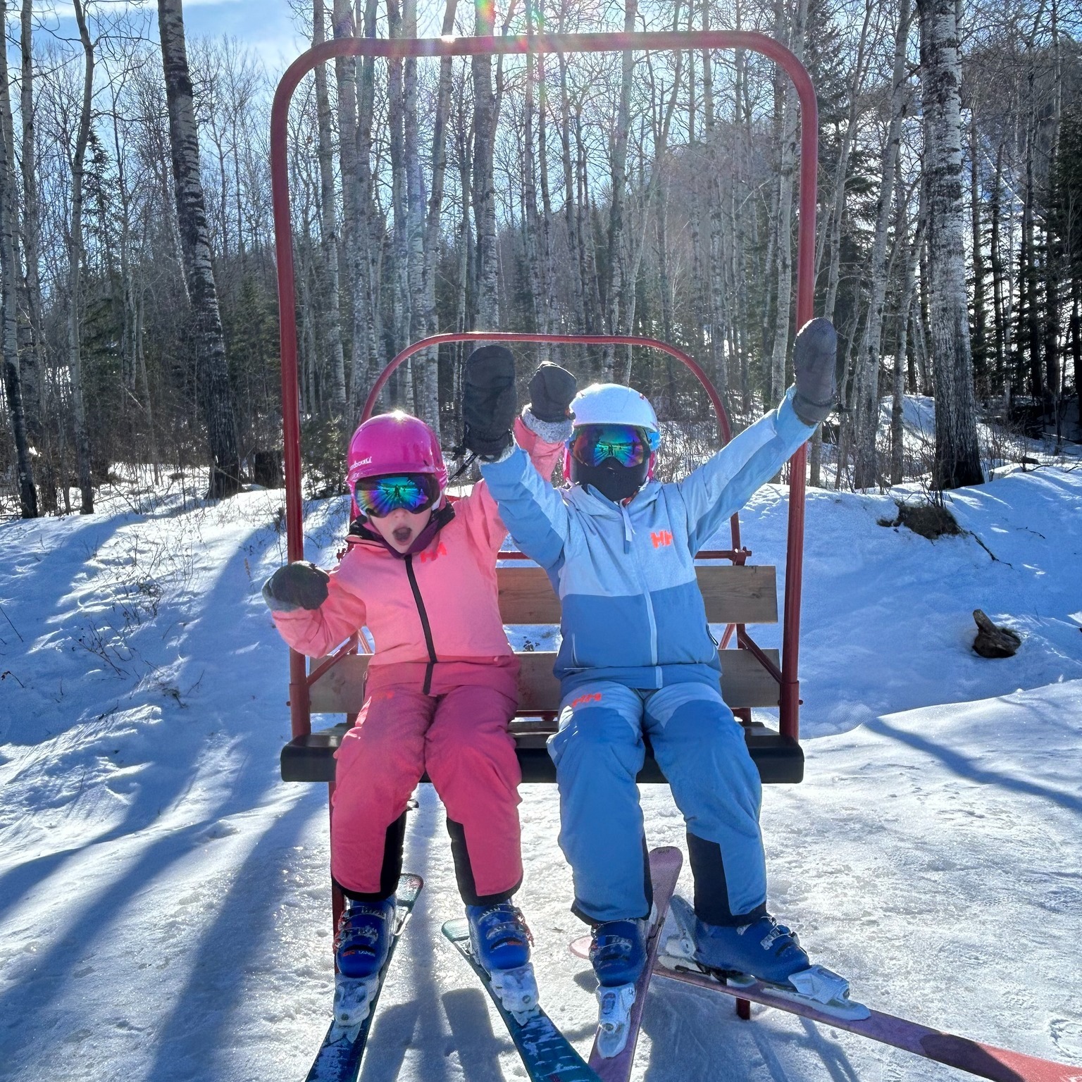 two cheering kids with arms raised, sitting on a ski lift above a treed ski hill on a sunny winter day.