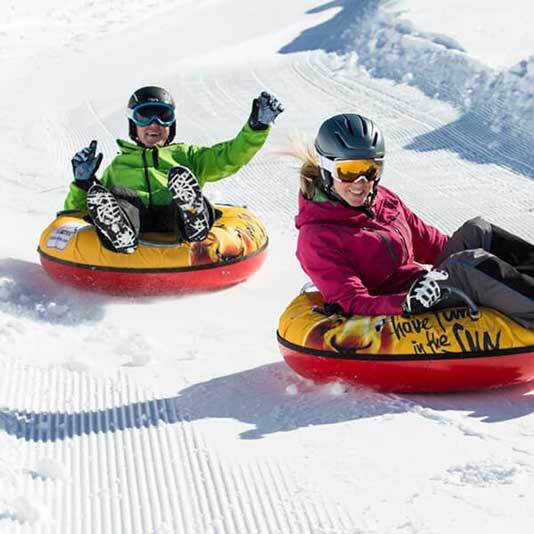 two kids wearing snowsuits and helmets, smiling widely as they ride inflatable tubes down a trail on Mount Baldy on a sunny day