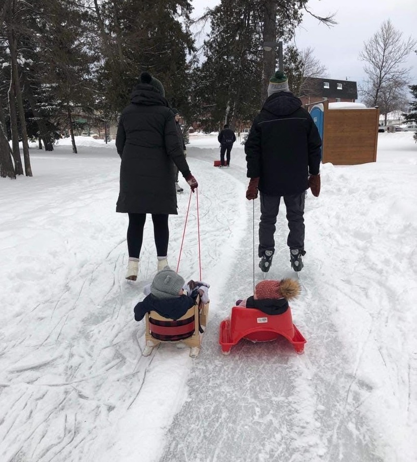 two people skating on an outdoor ice trail, each pulling a baby in a sled behind them. 