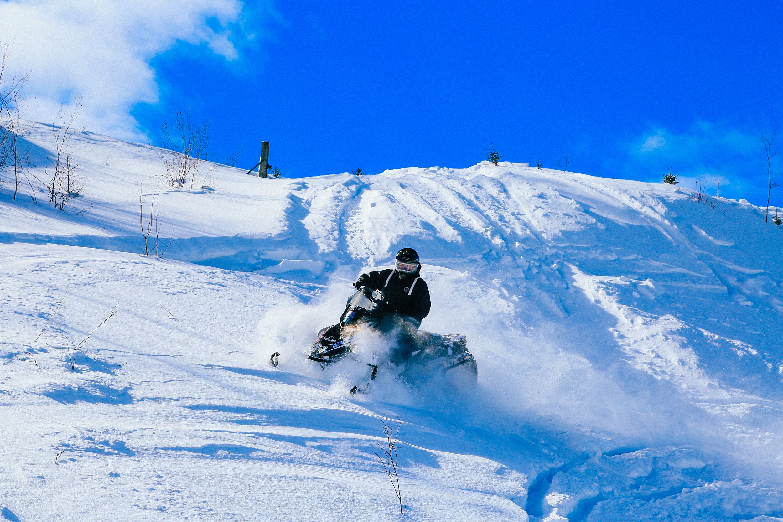 A rider on a snowmobile crossing a steep hill covered in soft powdery snow that flies up around it. The sky above is bright blue.