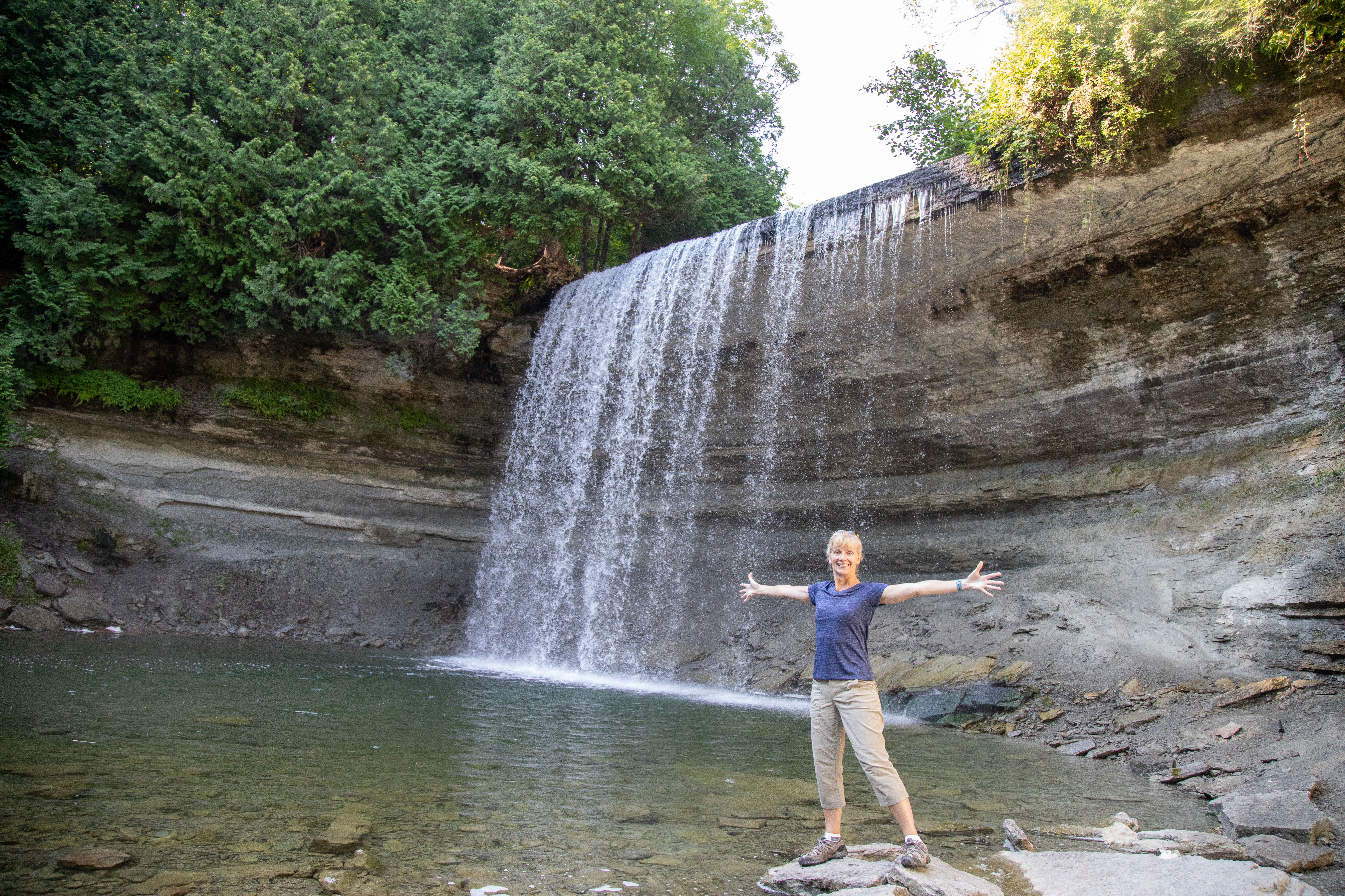 Bridal Veil Falls; a smiling woman holding her arms wide stands on a rock in front of a large waterfall off a steep rock cliff surrounded by lush green trees. It falls into a glassy green pool at her feet. 