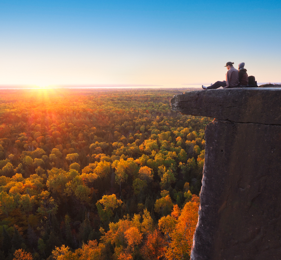 Cup and Saucer Trail; Two people sit at the top of an extremely high rocky outcropping, overlooking a treed valley under an orange sunset.