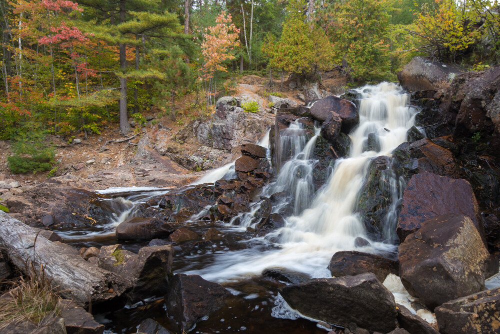 Duchesnay Falls; a rocky waterfall surrounded by thick green forest, the leaves slightly turning to red for autumn. 