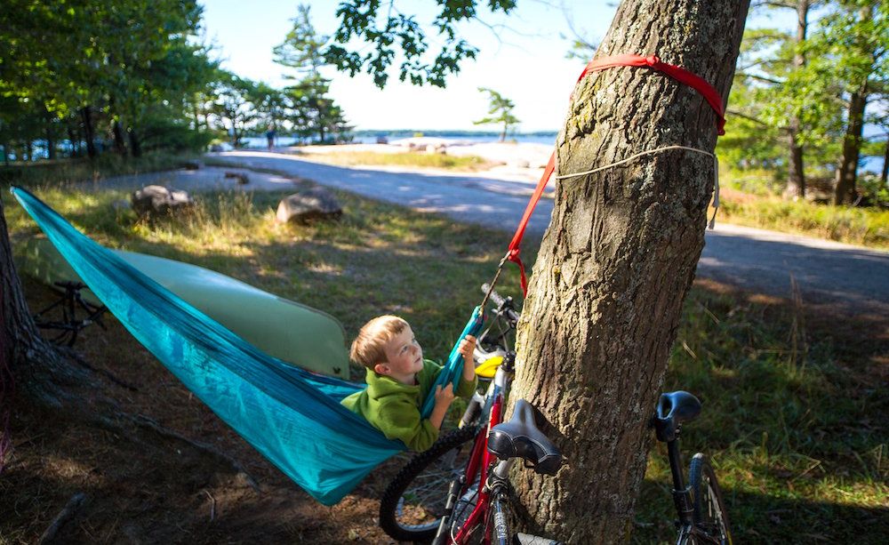 A boy laying in a hammock strung between two trees, looking upward. His bicycle is propped up next to the hammock and there is forest and a campground road in the background.