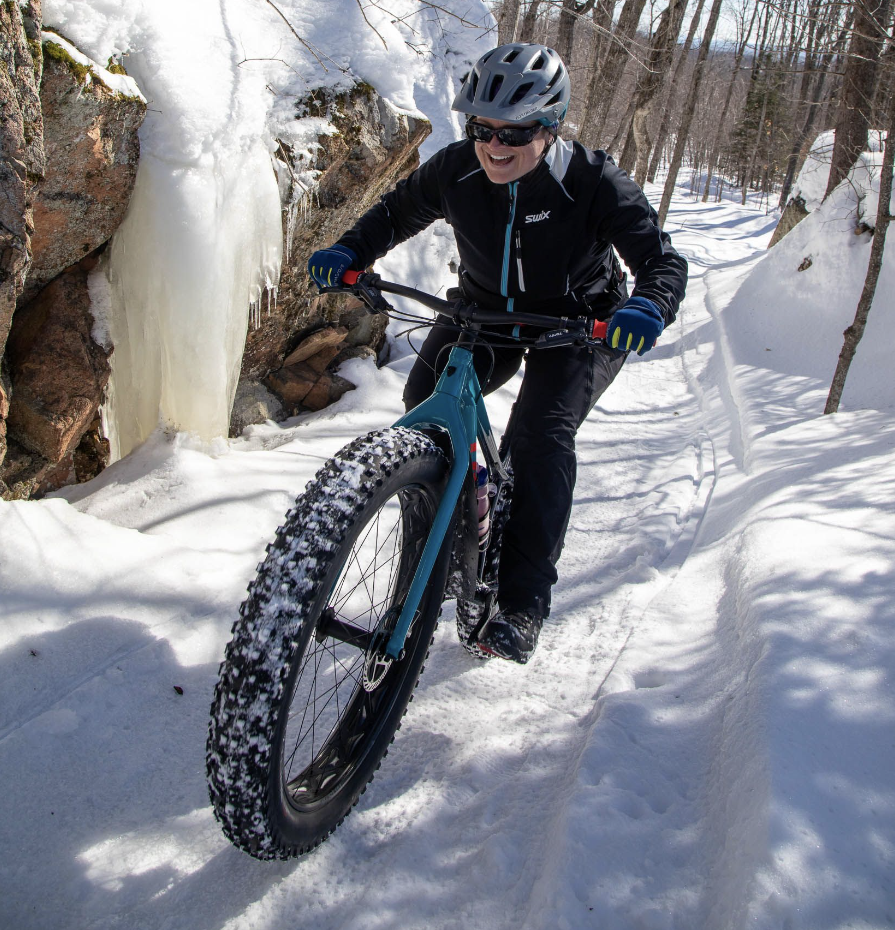 A smiling fatbiker riding through a snowy forest on a sunny day.
