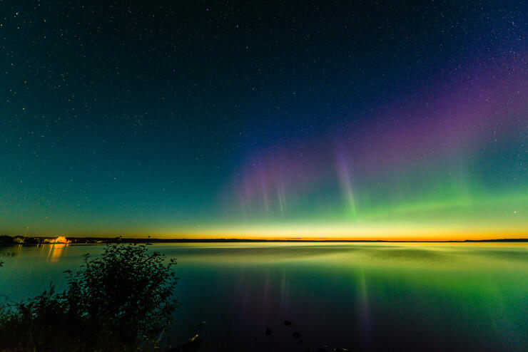 A stripe of aurora borealis lights up a dark night sky with brilliant purple, green, and yellow tones that are reflected in a glassy still lake below. A lit up cabin is in the distance.  