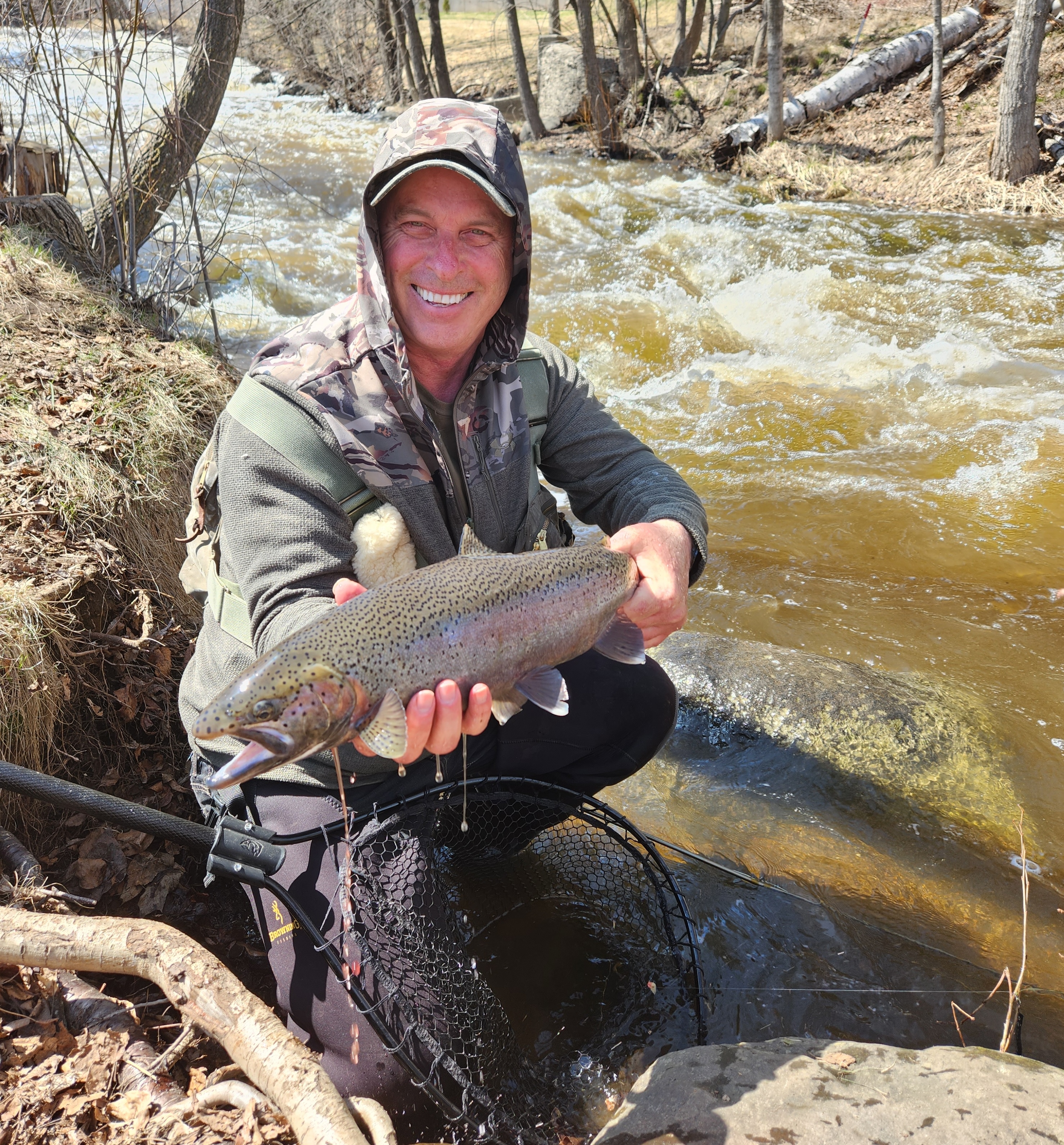 A smiling man crouching in a rocky, rushing stream, holding a large fish.