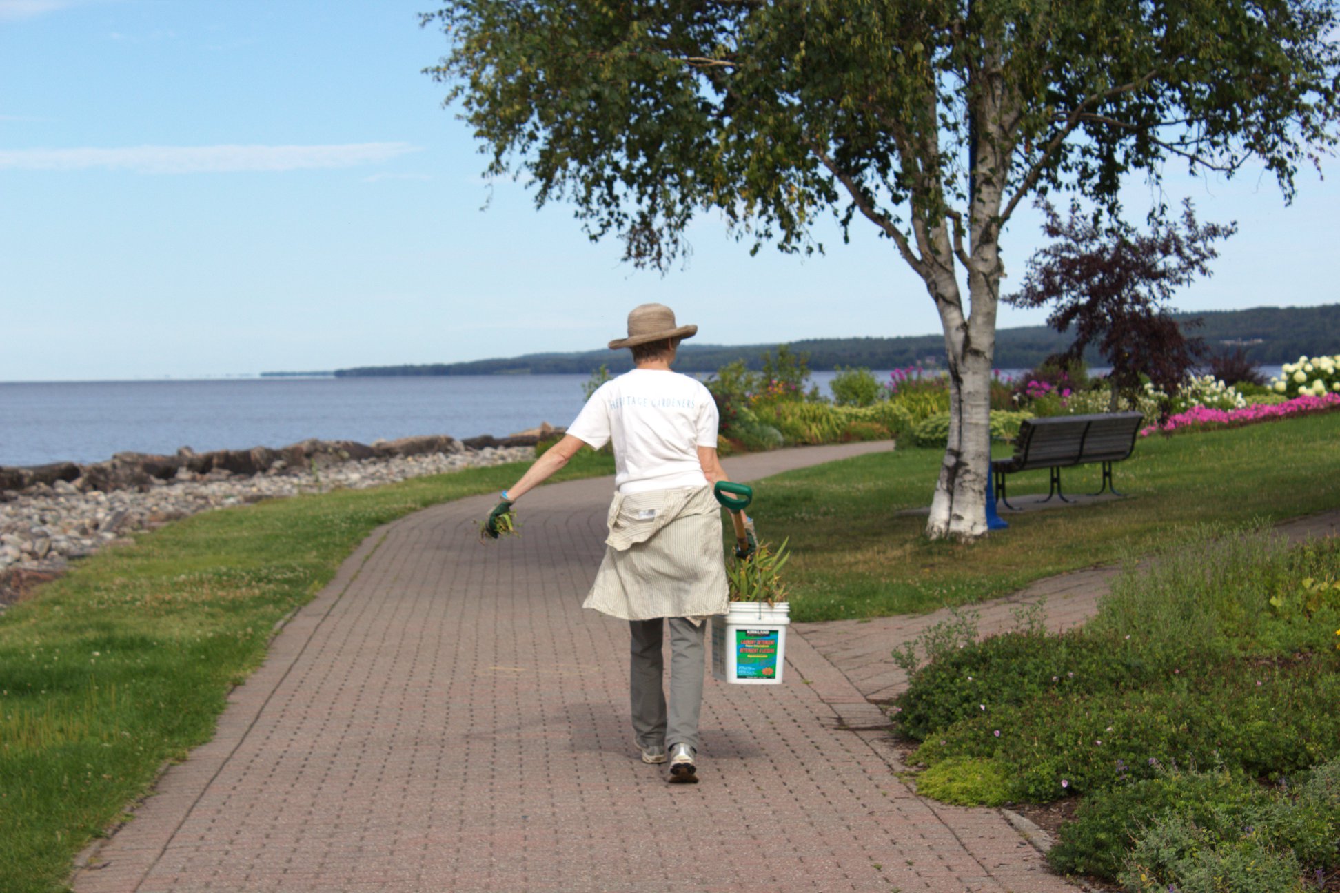 A person wearing gloves. a sunhat and carrying a bucket, walking down a brick walkway in a beautiful waterfront garden under a blue sky.