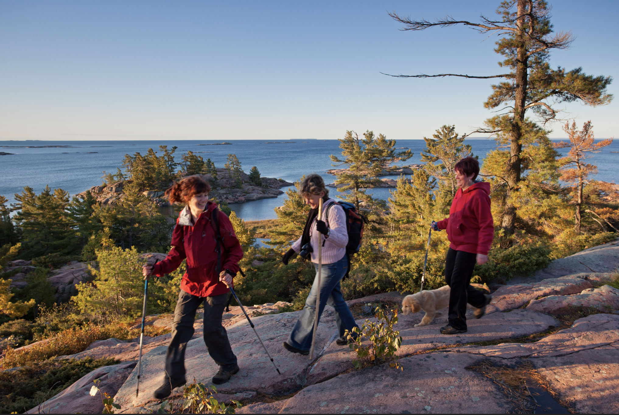 3 smiling people hiking down a tree-covered rock bank in front of a large lake in Killarney Provincial Park.