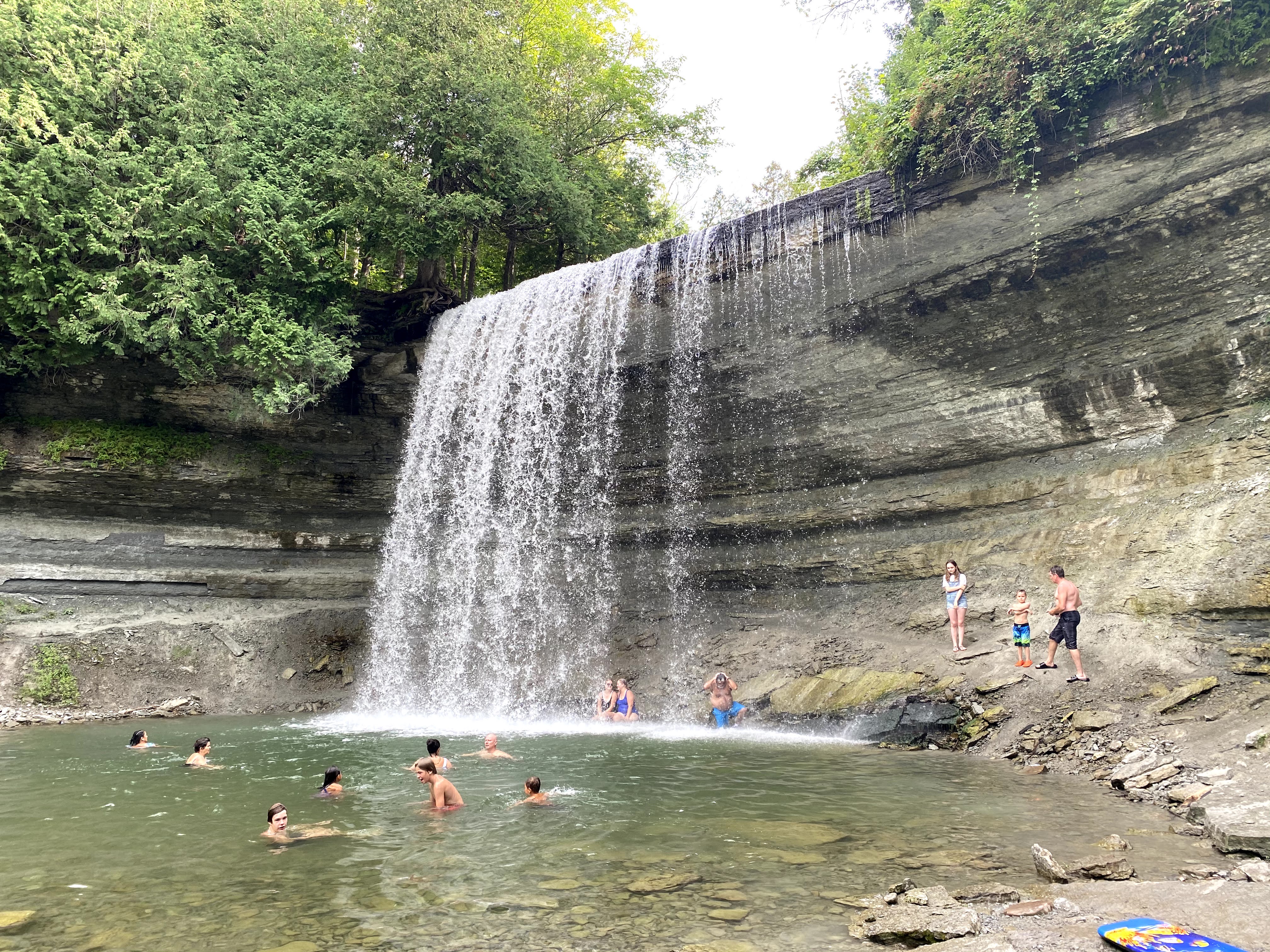 Bridal Veil Falls; a wide waterfall that pours over a treed rock cliff into a still, clear pool at the its foot. There are several bathers swimming in the pool. 