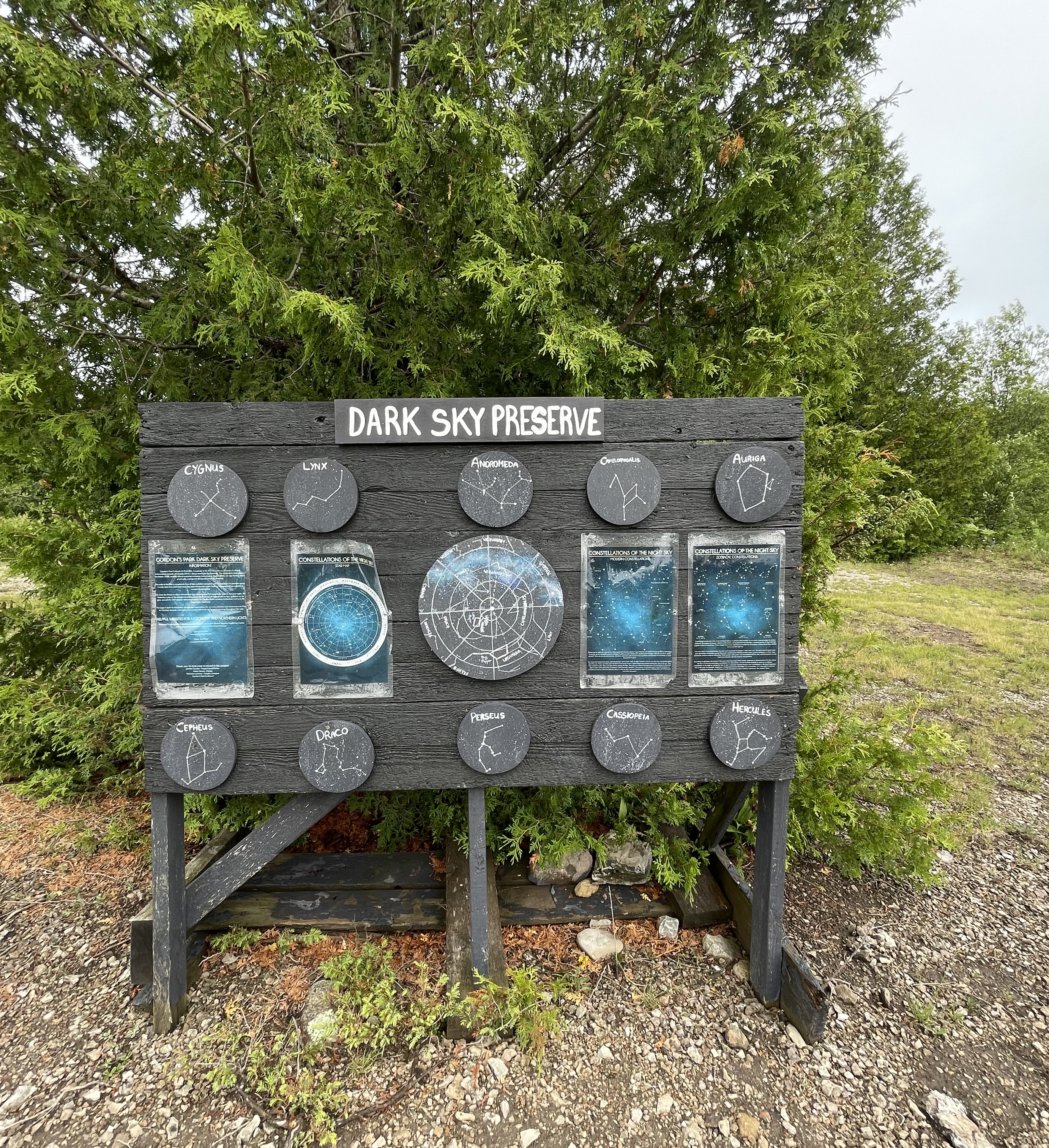 A wooden informational sign surrounded by green bushes that reads "Dark Sky Preserve" and shows visitors various constellations that they could look for. 