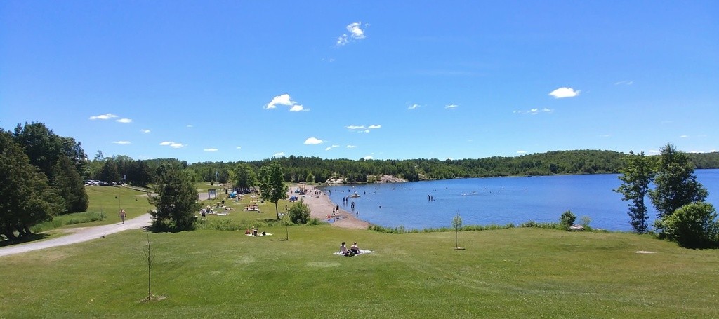  a sandy beach surrounded by green grass and trees, along a large, very blue lake under a blue sky. The beach is dotted with people.
