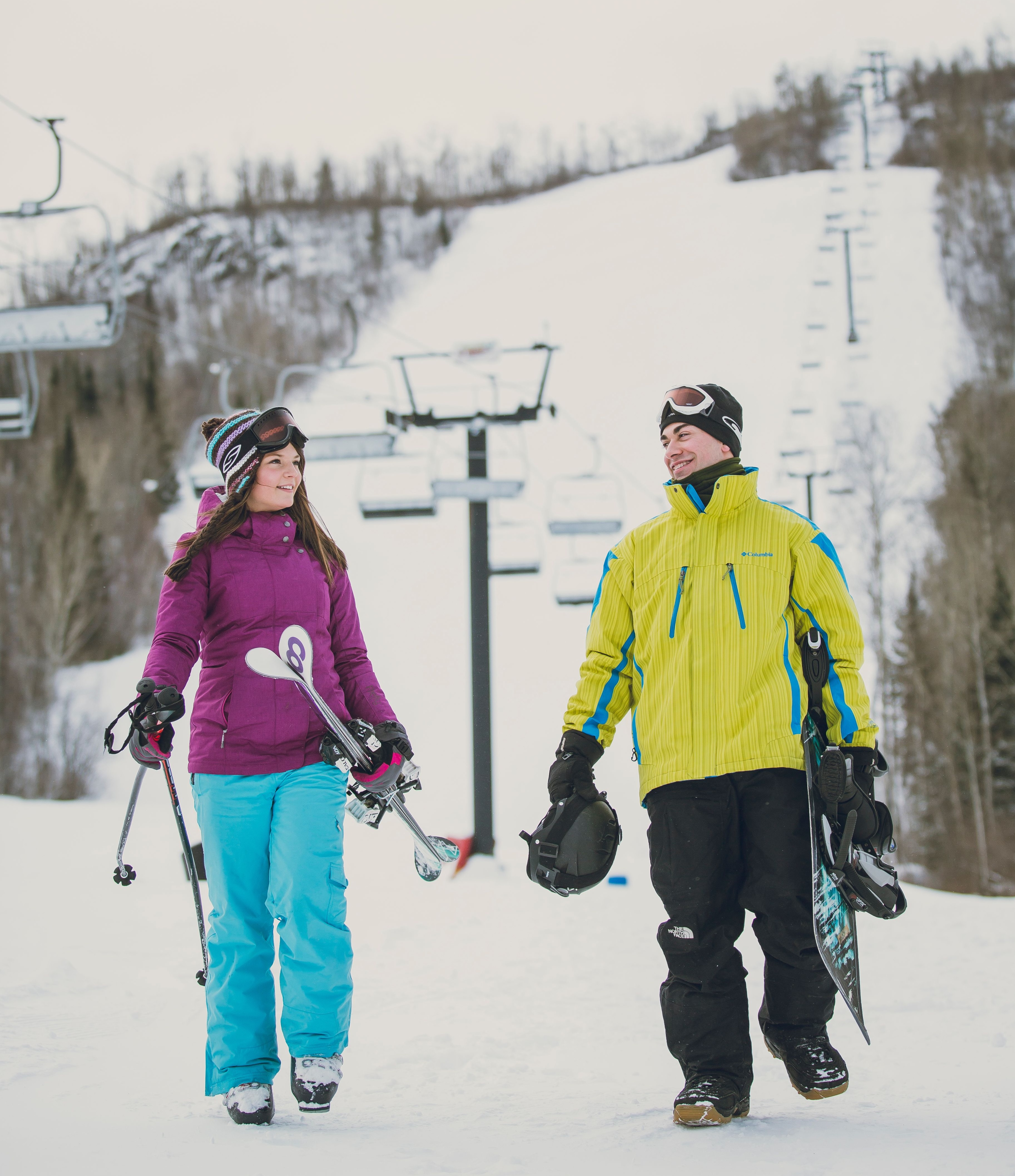A skier and snowboarder talking and smiling as they walk under a ski lift at the bottom of a ski hill.