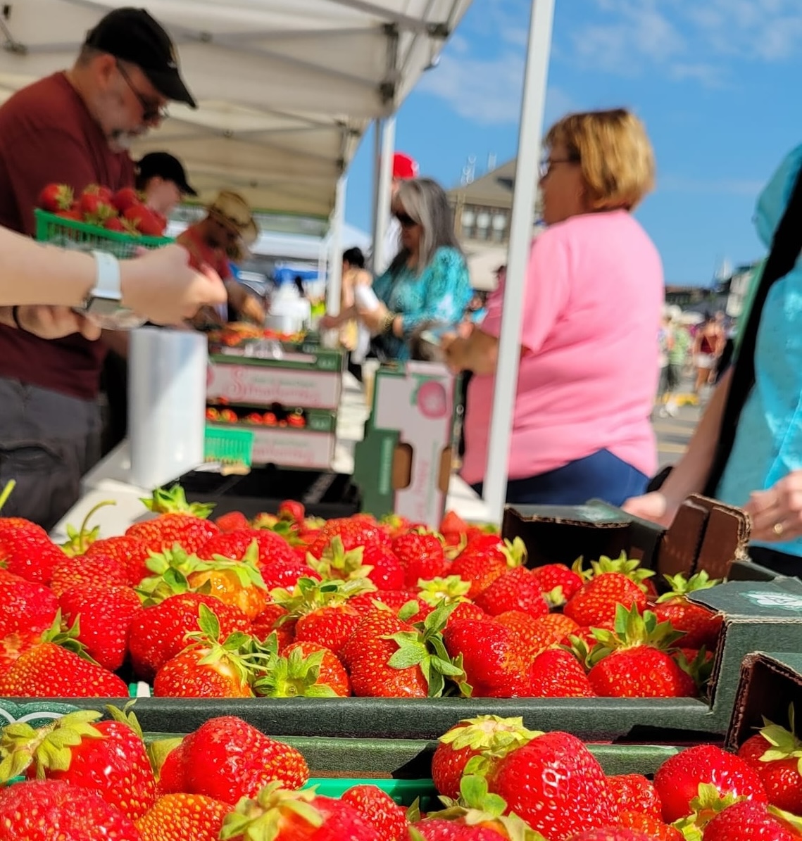 Baskets of red strawberries on a table at a farmers' market, shoppers paying for their produce in the background.