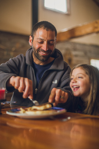 a man and young girl sitting a a table. The smiling man is cutting up some pancakes on a plate and the girl, sitting next to him, is laughing. 