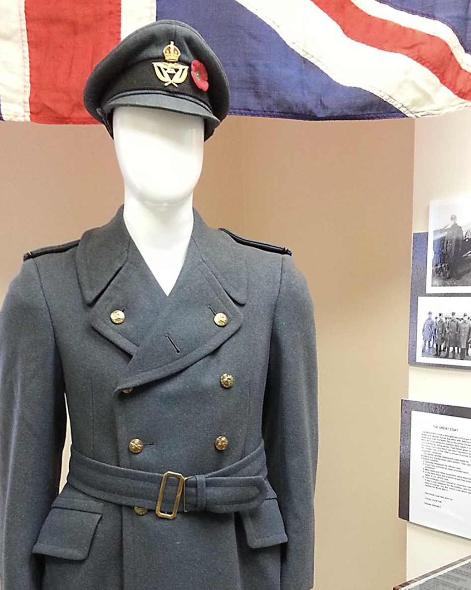 A World War 2 era air force coat and hat on a mannequin, in front of a flag and a series of black and white photos on display at the Northwestern Ontario Aviation Heritage Centre.