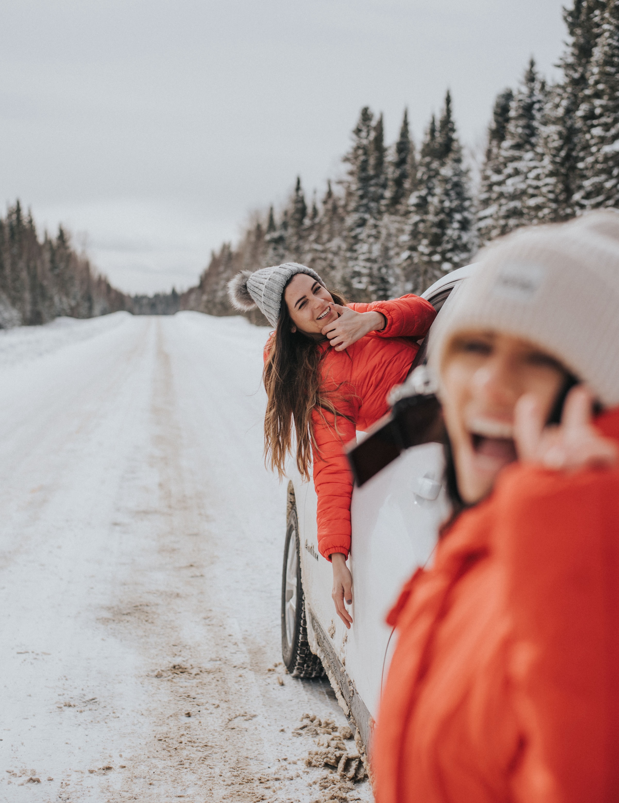two people in winter outdoor clothing smiling and laughing while posing with a white car on a remote snowy road surrounded by snowcovered forest.