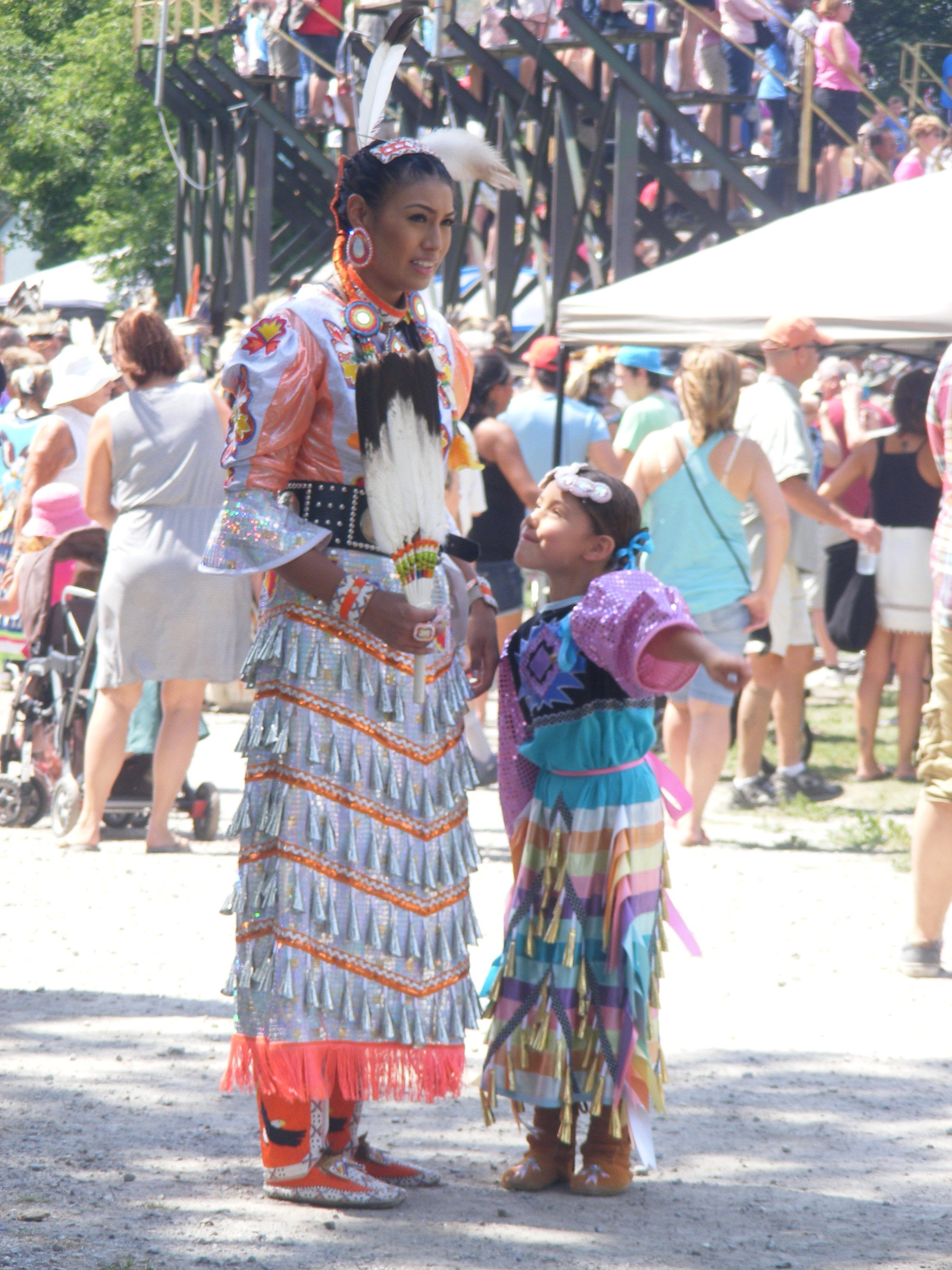 An indigenous woman and child in traditional dancing clothes at a pow pow on a sunny day. The child is pointing and speaking to the woman, who is smiling. Grandstands and a crowd are in the background.