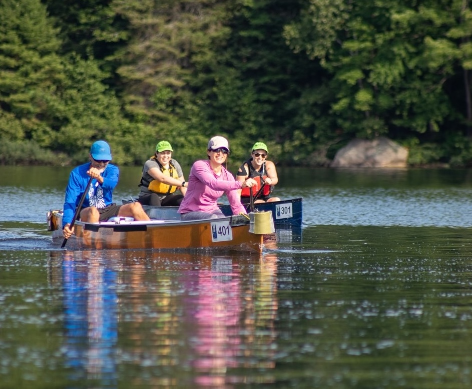 A group of smiling paddlers in canoes with race numbers attached to their bows, paddling down a glassy river in front of green forest on a sunny day.  