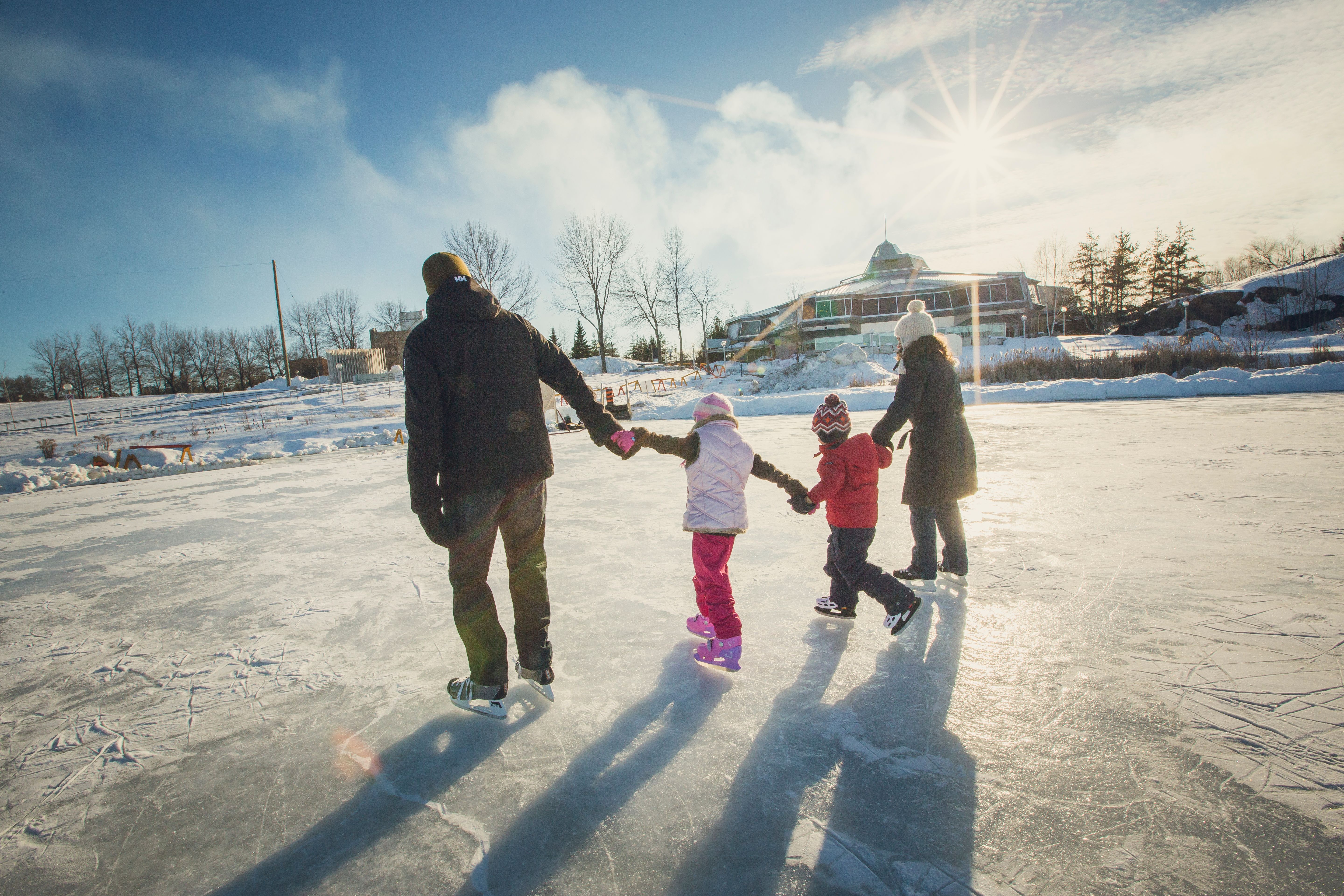 Two adults and two children hold hands in a line as they skate across a frozen pond in late afternoon on a sunny winter day. Their shadows stretch long behind them. 