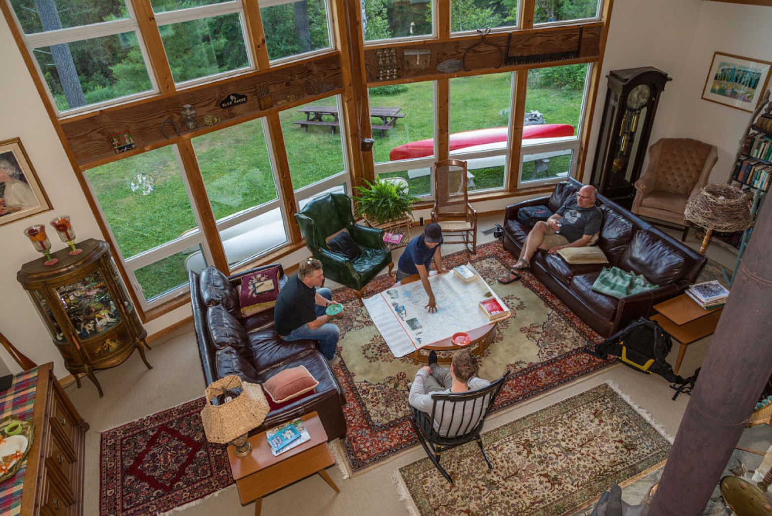A high shot looking down on a family in a large cabin sitting room, sitting on couches around a coffee table. There are tall windows that fill one wall, and outside there is green grass, forest and canoes.