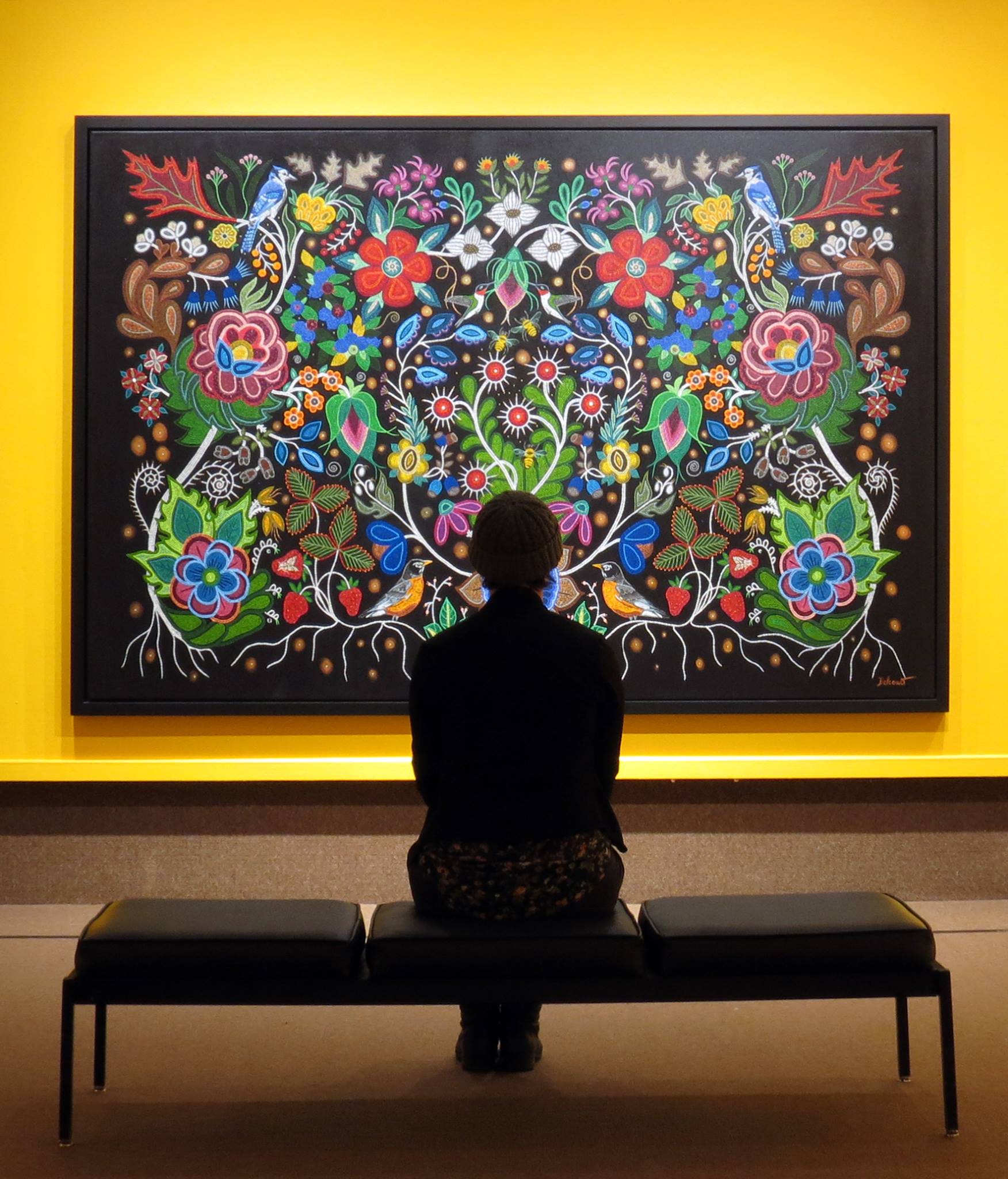A silhouetted person sitting on a bench in front of a large painting of flowers on a black background, hung on a yellow gallery wall.