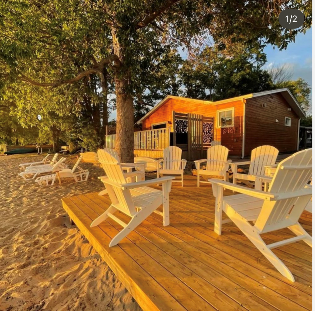 a sandy beach at sunset, with green trees, a small wooden cabin, a wooden deck, and white wooden chairs  on it.