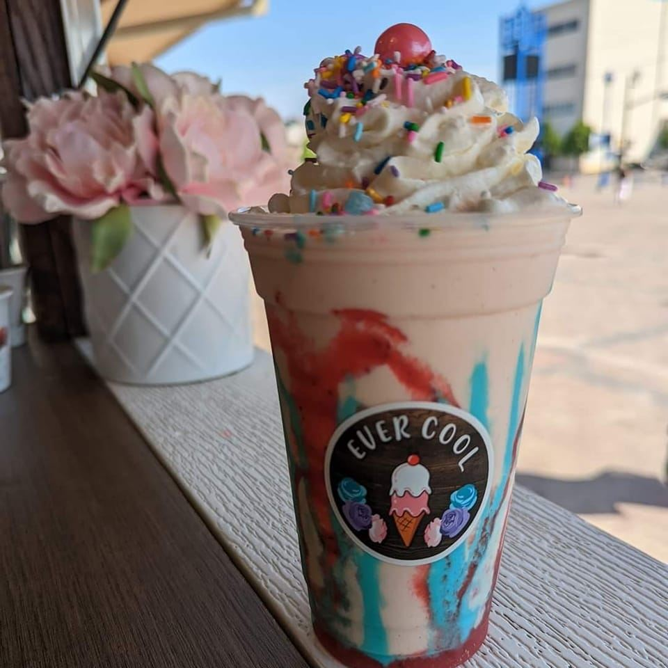 a colourful milkshake in a glass that reads "Ever Cool", covered in whipped cream, sprinkles and a cherry. It is sitting on a window ledge next to a pot of pink flowers on a sunny day. 