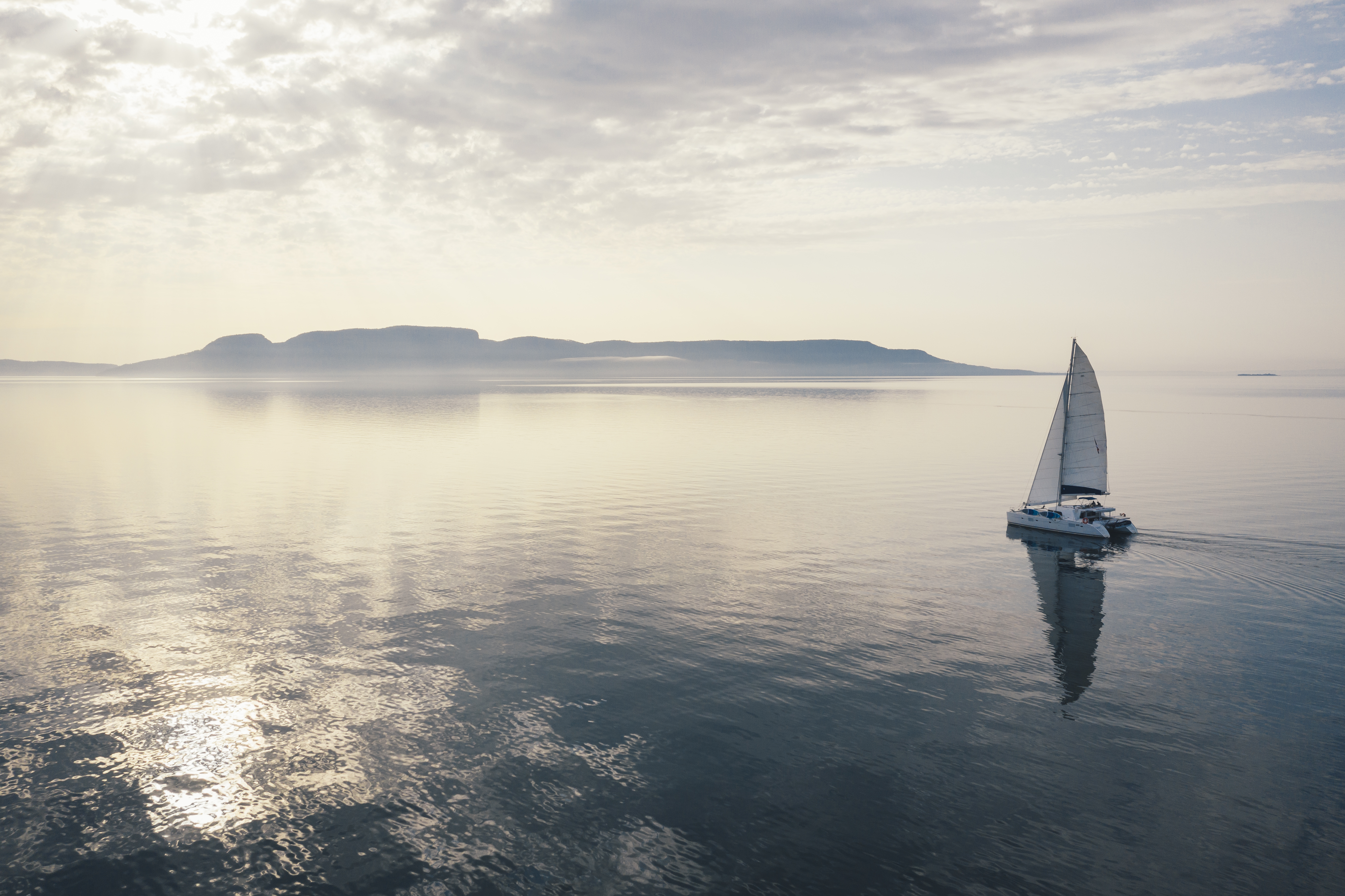 A sailboat sailing into golden sunlight, toward Sleeping Giant on the misty horizon.