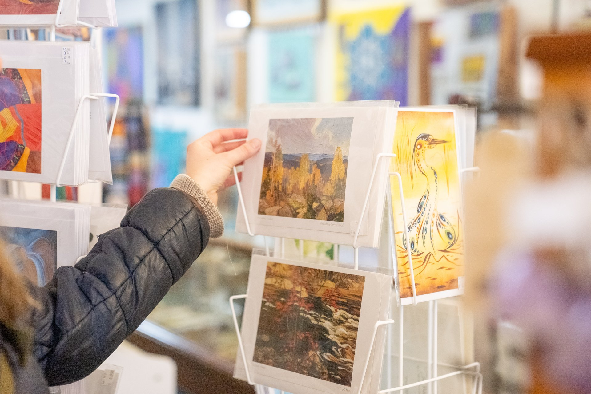 The Ahnisnabae Art Gallery; a shopper's hand picks up an art print off a rack. The background is full of many more racks of colourful art prints.