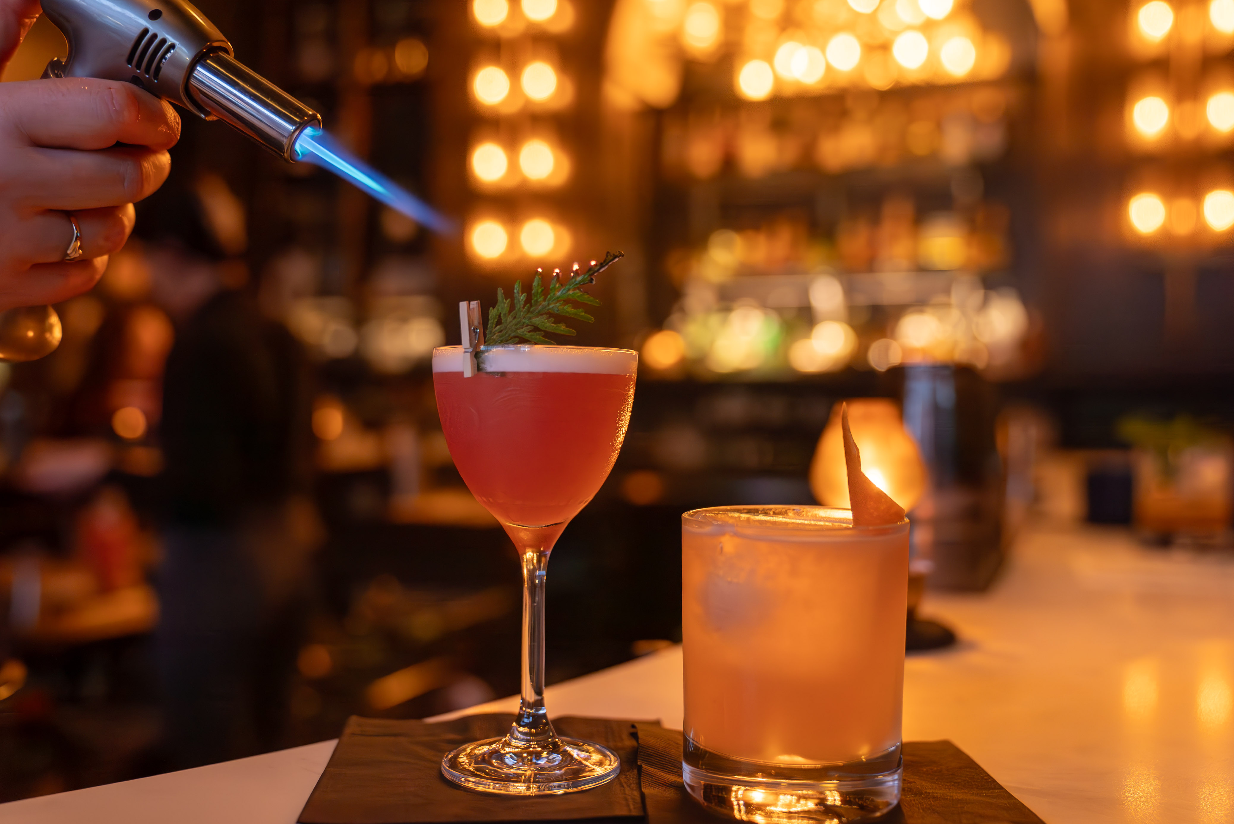 a hand bringing a lit kitchen torch toward two beautifully decorated cocktails in a dim lounge, warmly lit by a wall dotted with lights that's in the background. 