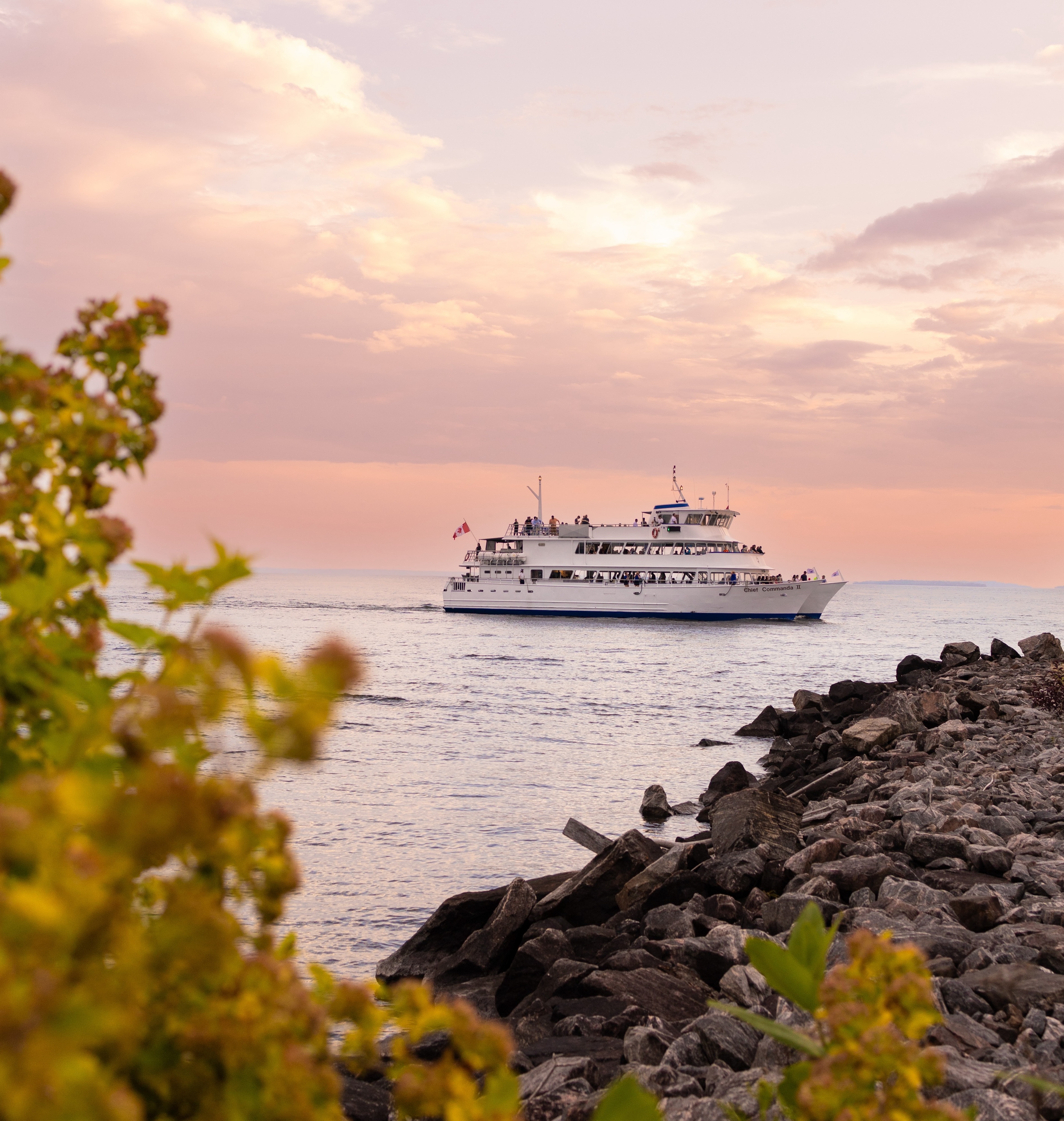 The Chief Commanda 2; a large passenger cruise ship sailing in the distance on Lake Nipissing, the sky beautifully lit by a delicate pink sunset. Green foliage and a rocky shore are in the foreground.