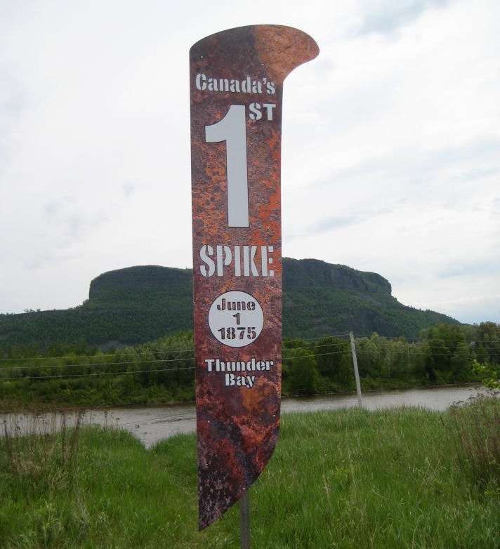 The marker for the First Spike in Thunder Bay; a large metal sign shaped like a railroad spike with the words "Canada's First Spike June 1, 1875 Thunder Bay" on it, surrounded by greenery with Mount McKay in the background. 