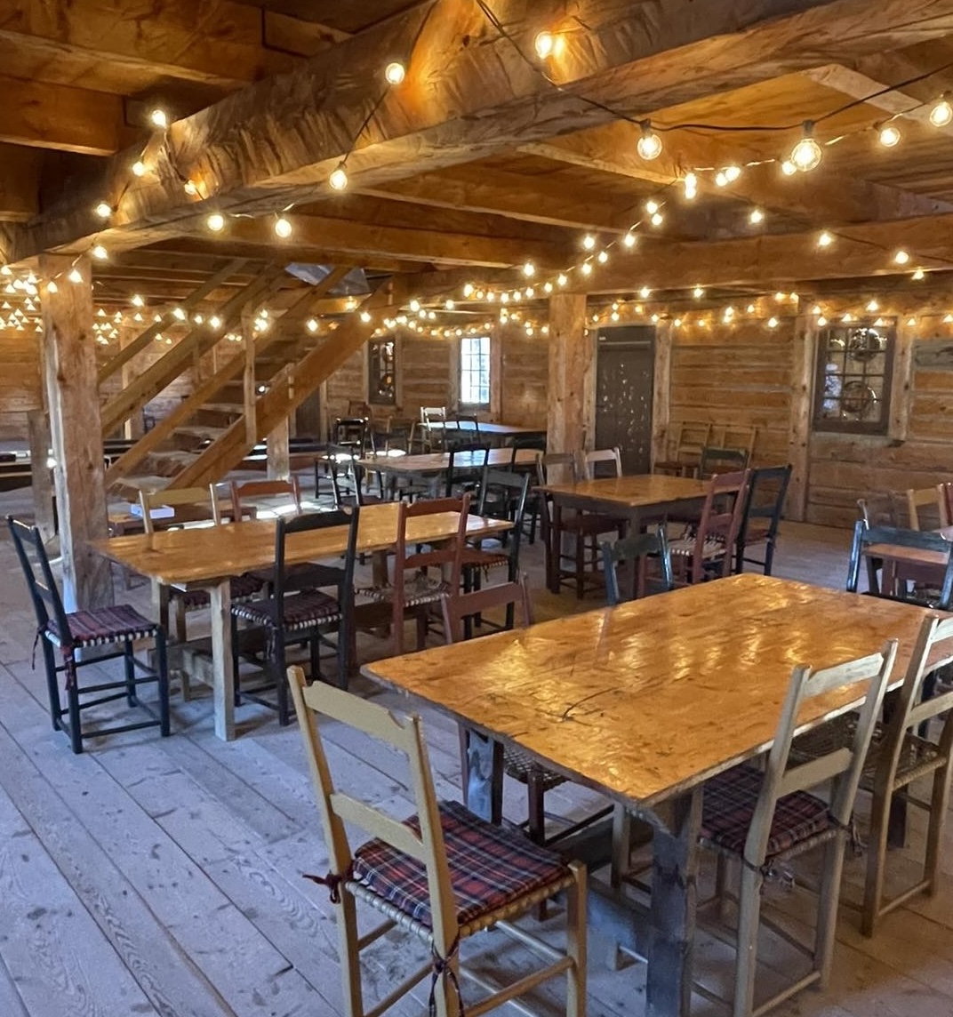 a banquet hall at Fort William Historical Park; several wooden chairs and tables in a large room with rough wooden floors, walls and rafters. There are strings of white lights adorning the rafters and giving soft light to the room.