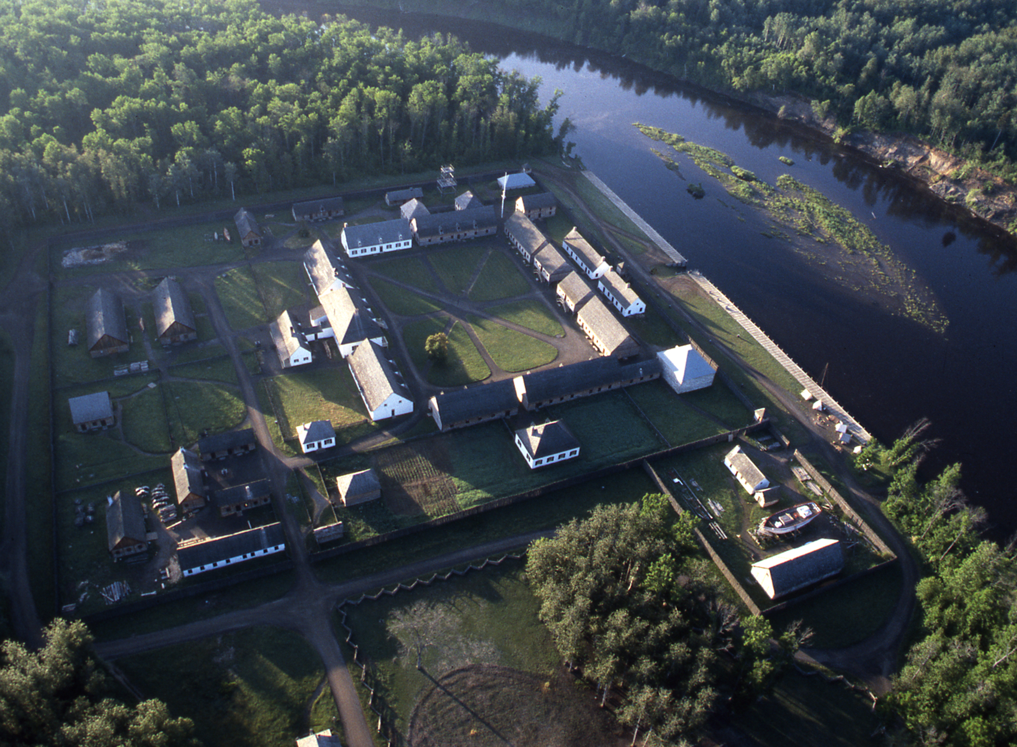 an aerial shot of Fort William Historical Park; several large, well-kept antique buildings arranged in a square layout on a grassy lawn which makes up the whole of the park. It is surrounded by thick green forest and a wide, calm river on side.