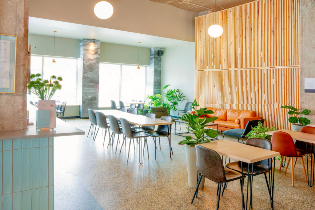 The Woodside Bar at Goods & Co, Thunder Bay; a bright, fresh-looking room with several long wooden tables with chairs, large potted plants, light wood slats on some walls and light blue tile on others.