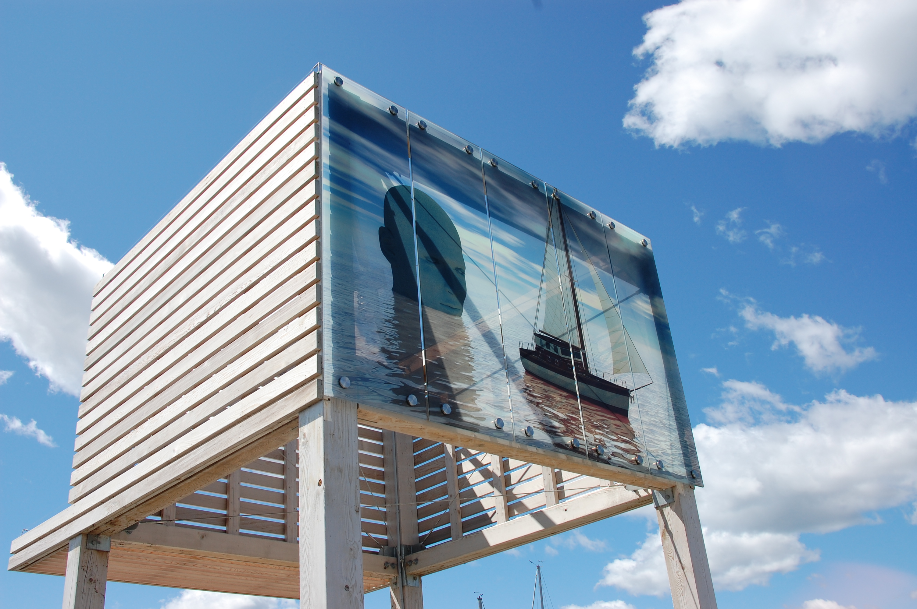 "Ulysses" by Mark Nisenholt, Lantern Art at Prince Arthur's Landing; a large wooden slat box set on high vertical beams, with the blue sky in the background. On the front of the box is a painting of a sailboat on the bay with the silhouette of a large human head rising out of the water. 