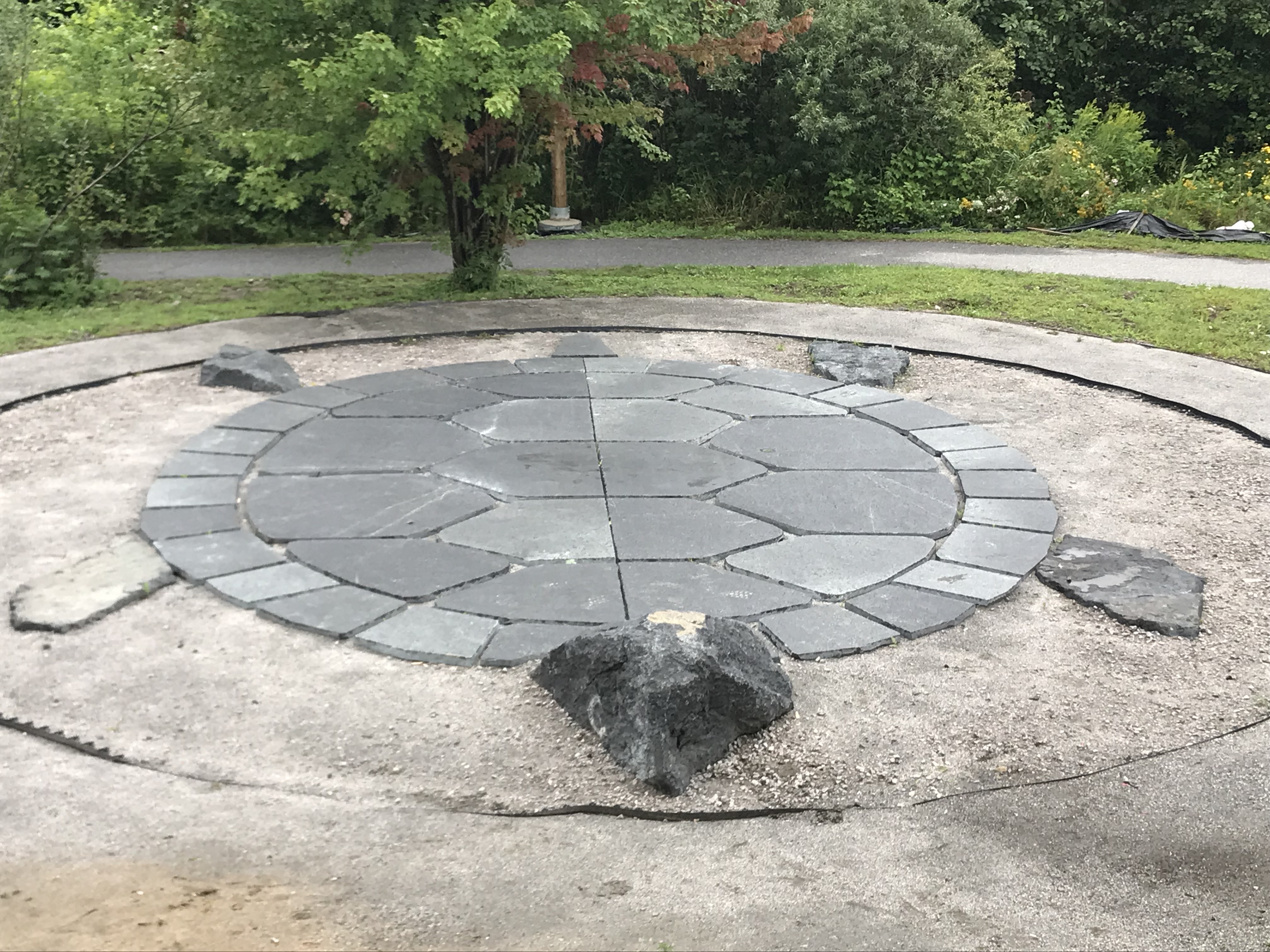 Miskwaadesi; a sculpture of a turtle made of flat stones inside a circle, laid in a greenspace surrounded by trees and a walking path. 