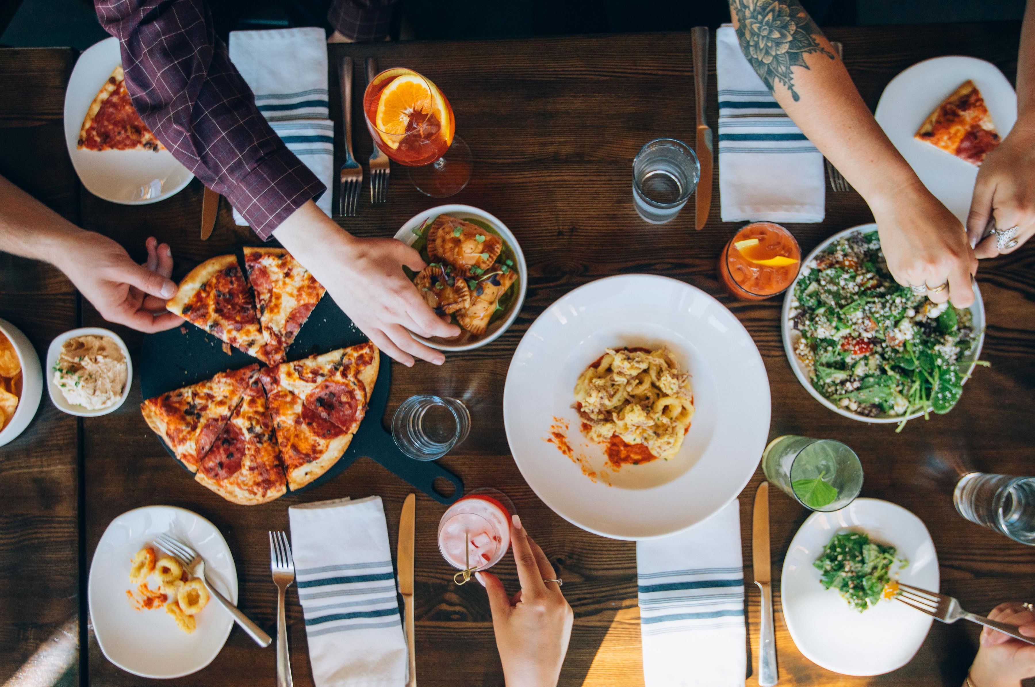 A high shot looking down on a table spread with pizza, salads, beverages and plates, while diners' hands reach from all sides, picking up the food. 