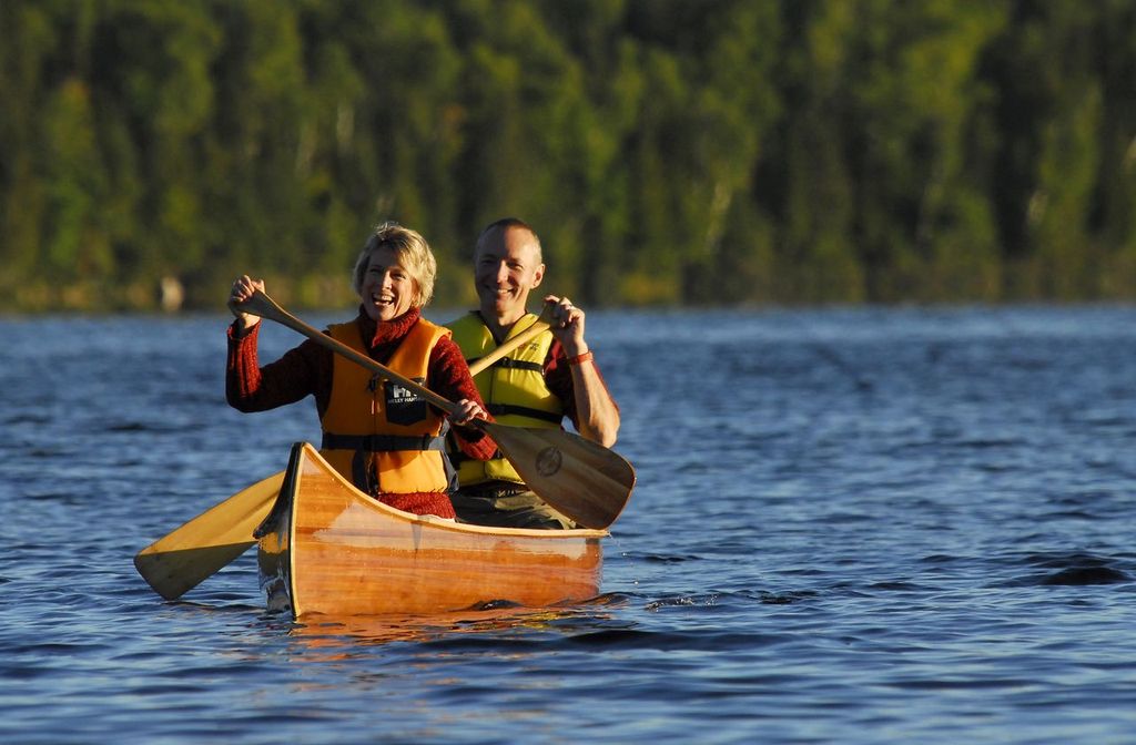 a smiling older couple paddling a canoe on a blue lake surrounded by green forest.