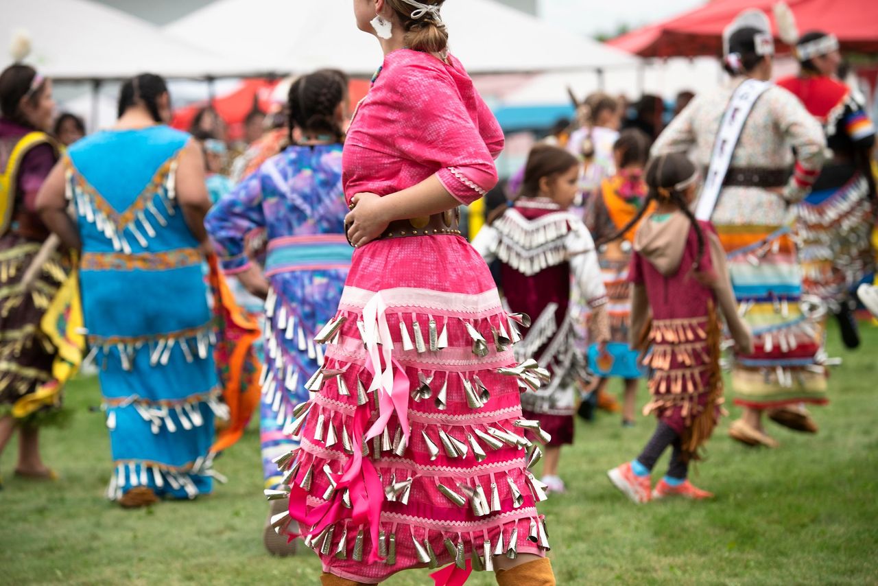 a group of dancers in colourful jingle dresses dance on green grassy pow wow grounds. The roofs of vendor tents are in the background.