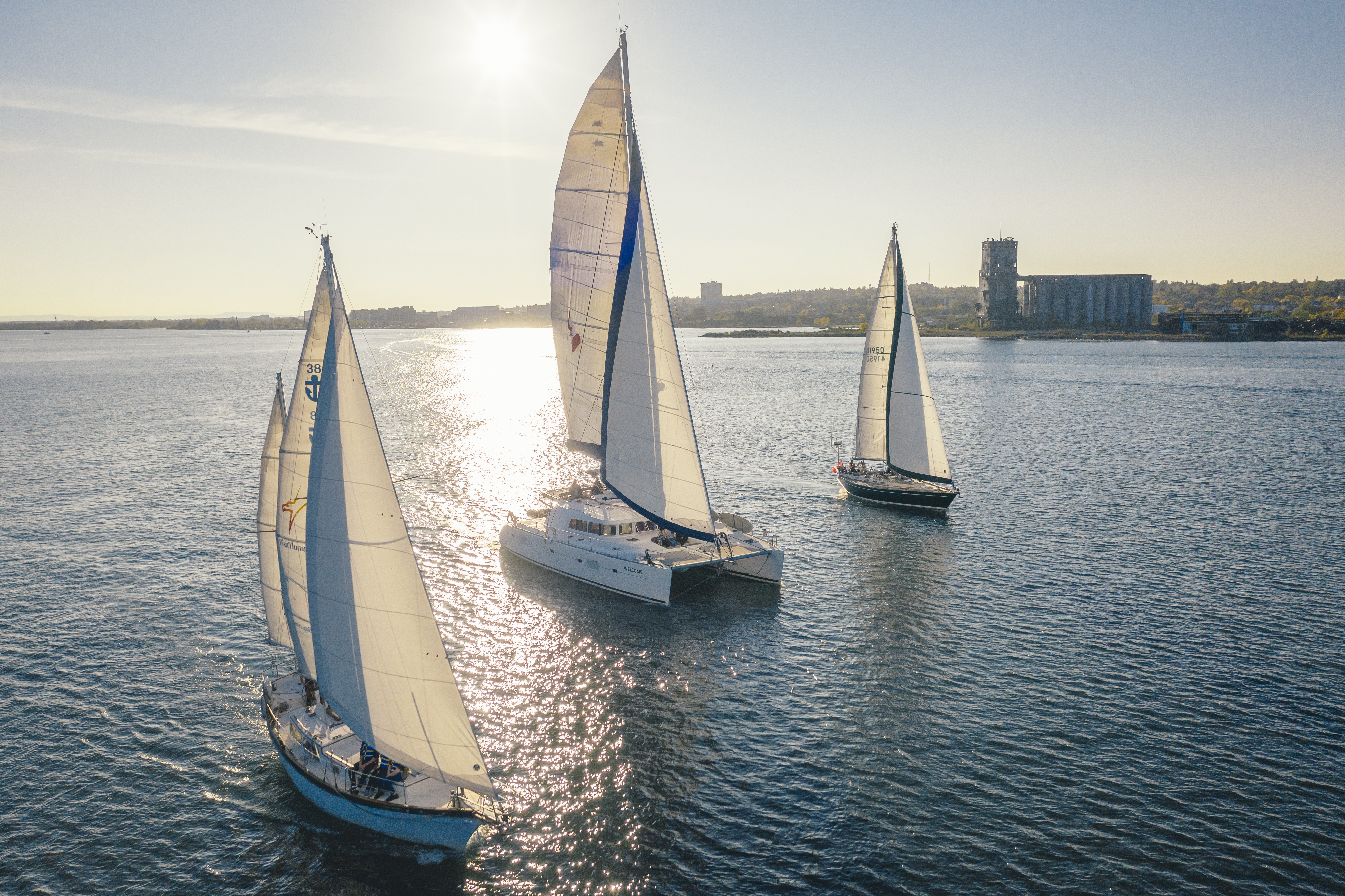 a fleet of 3 sailboats on the water with a high, bright sun shining through their sails. The port of the city of Thunder Bay is on the horizon. 