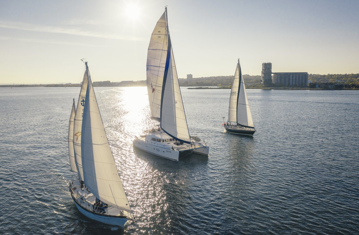 large white sailboats and a catamaran sailing on Lake Superior in late afternoon with the city of Thunder Bay on the horizon. 