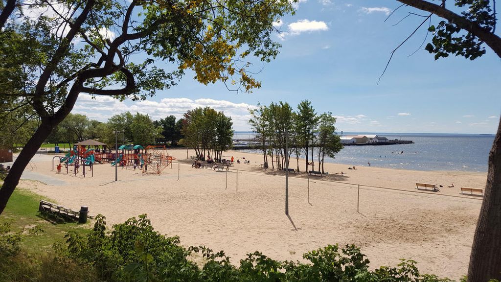 Shabogesic Beach; a large white sand beach with a playground structure, next to the large blue expanse of Lake Nipissing. Green foliage is in the foreground.