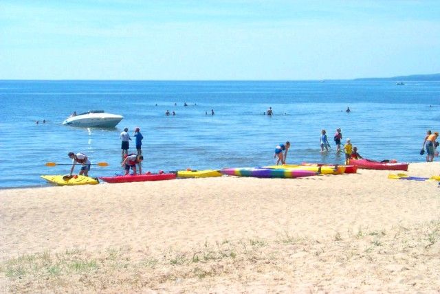 Shabogesic Beach, North Bay; a group of people with their kayaks lined up along the edge of a white sandy beach in front of a very blue lake on a clear, sunny day. There are many swimmers in the water.